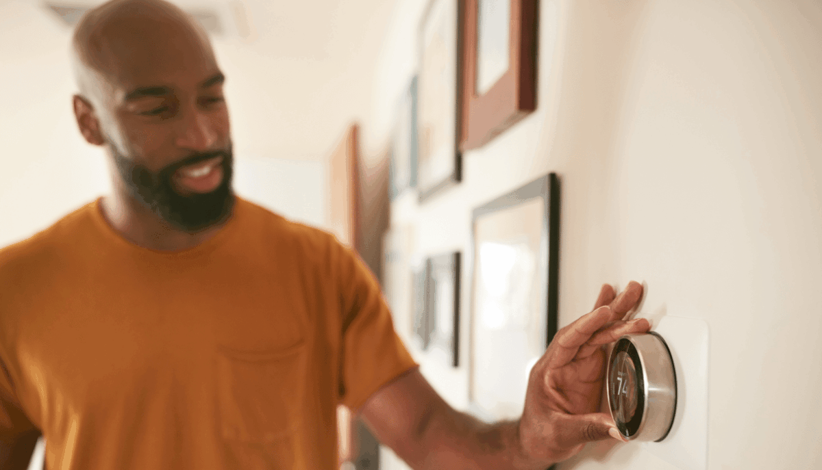 A man in an orange shirt reaching for his smart thermostat to adjust the temperature