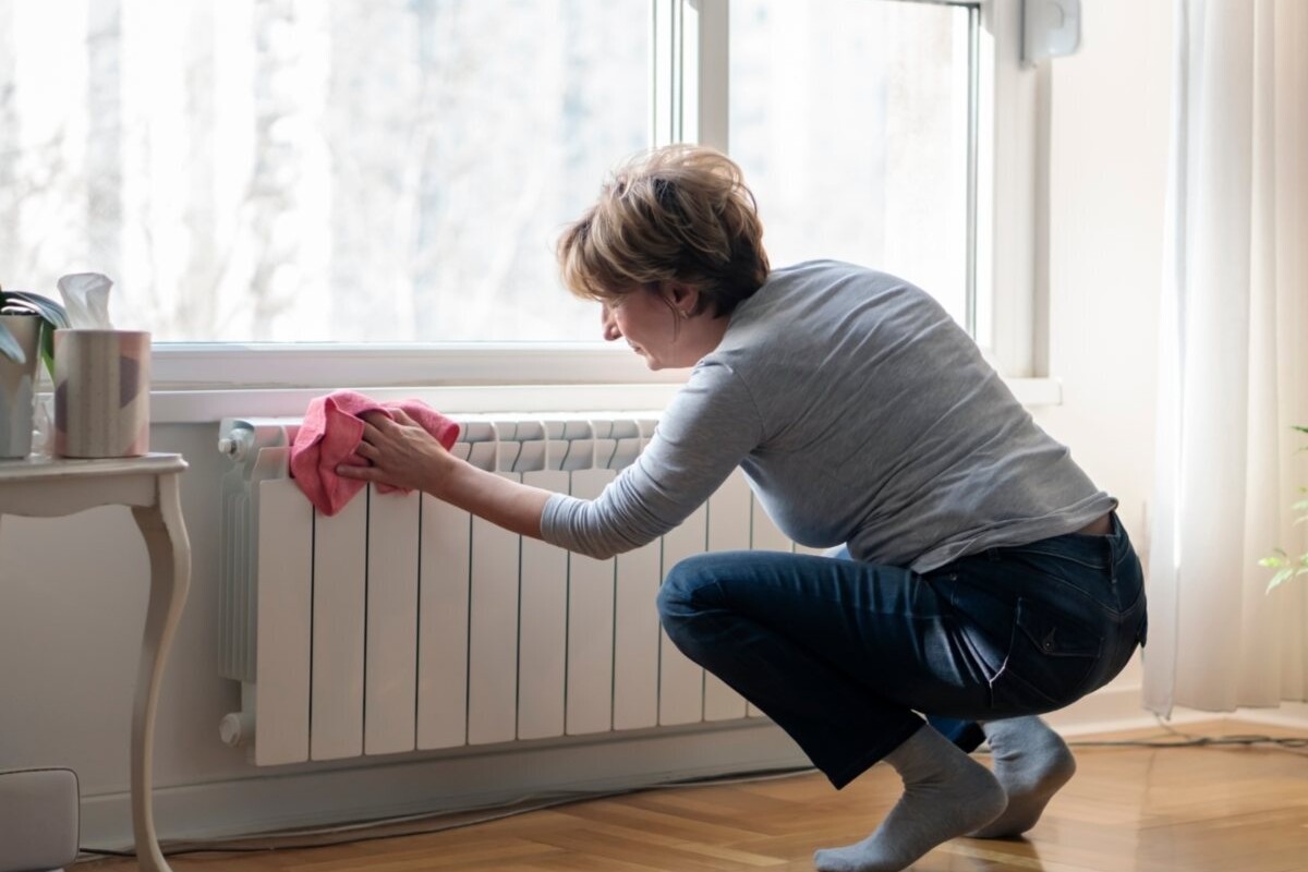 A woman crouches to brush dust off a heating unit in her home.