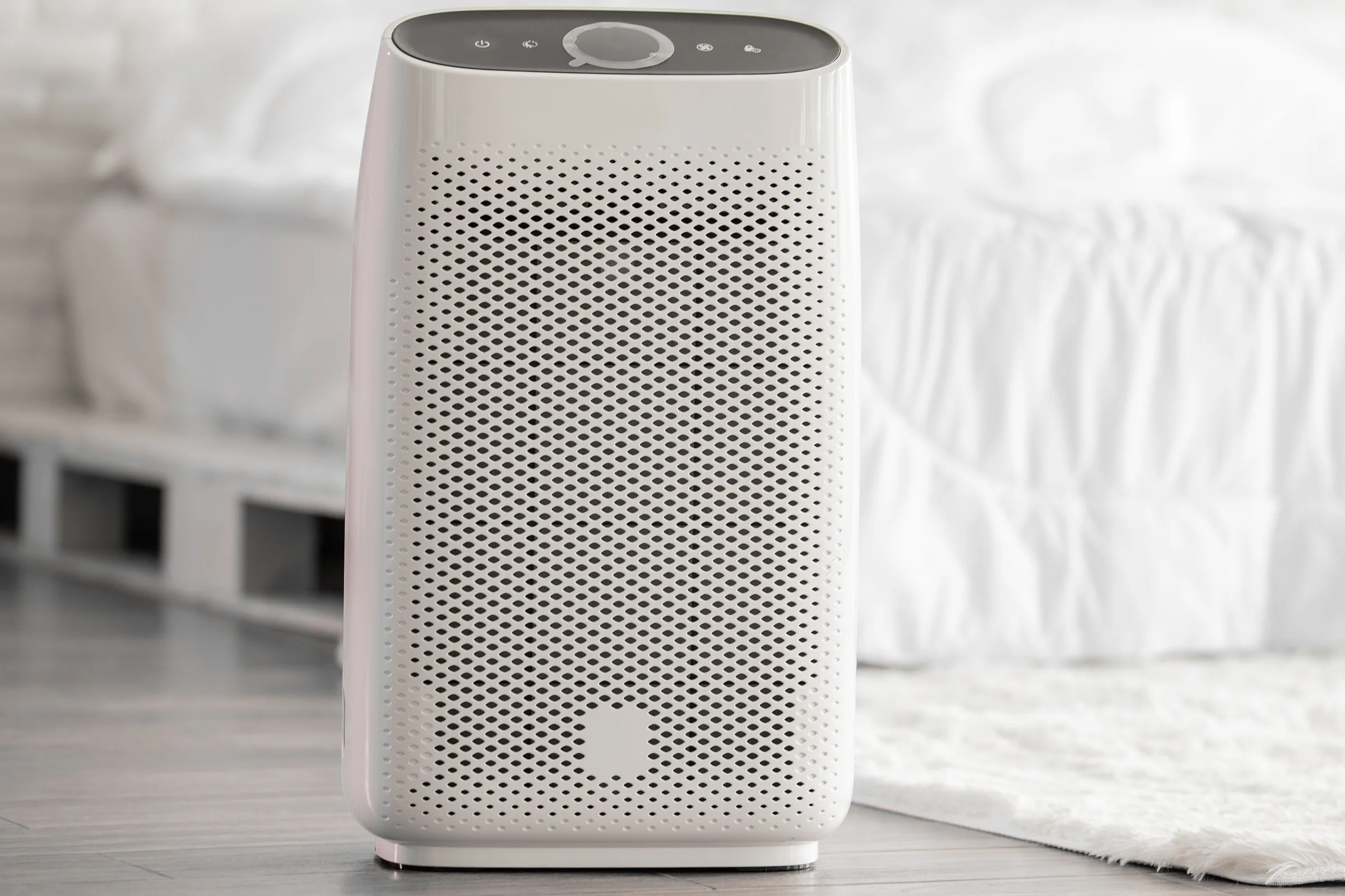 an air purifier on the ground of a white, modern room next to a bed.