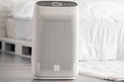 an air purifier on the ground of a white, modern room next to a bed.